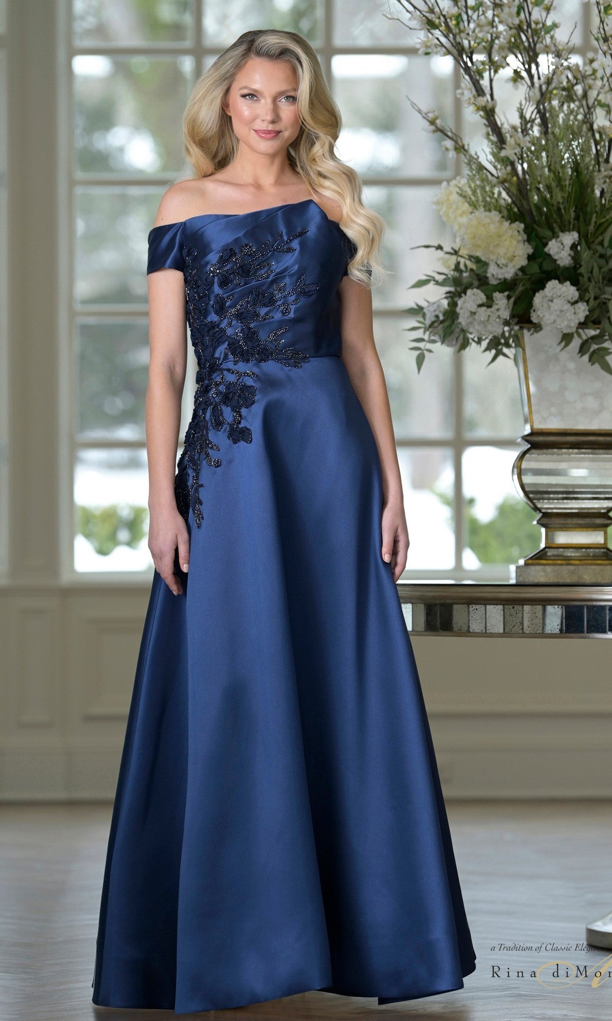 A woman stands indoors wearing the Rina Di Montella RD3501 long formal dress in light navy, featuring rich Mikado fabric, an off-the-shoulder neckline, black floral embroidery on the bodice, and a floor-length a-line skirt.